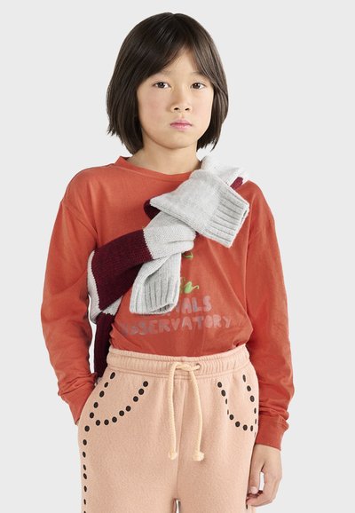 Camicia rossa a maniche lunghe con testo verde, pantaloni della tuta pesca con dettagli neri e tre sciarpe lavorate a maglia in grigio e bordeaux appese sulla spalla.