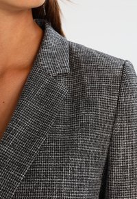 Blazer à carreaux gris et noir avec un col structuré, fabriqué en tissu texturé, offrant une coupe ajustée et des détails de couture subtils.