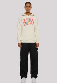 Crewneck hoodie in cream with a colorful "FROOT LOOPS" graphic. Features a front pocket and ribbed cuffs, paired with black pants and white sneakers.
