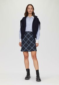 Marineblauw gebreid vest over een lichtblauw overhemd met knoopjes, gecombineerd met een geruite rok in marineblauw en lichtblauw, en zwarte enkellaarsjes.