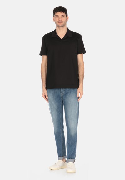Polo shirt nera con colletto aperto, abbinata a jeans azzurri chiari e sneaker bianche. Modello in piedi contro uno sfondo bianco semplice.
