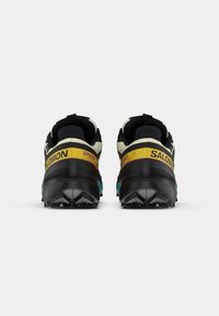 Baskets de trail running avec un design noir, jaune et turquoise. Elles présentent une semelle épaisse, un talon renforcé et une tige à motifs pour une meilleure adhérence.