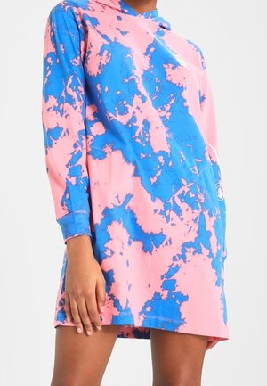 Femme portant une robe sweat à capuche à manches longues, tie-dye rose et bleu, coupe décontractée, debout devant un fond clair uni.