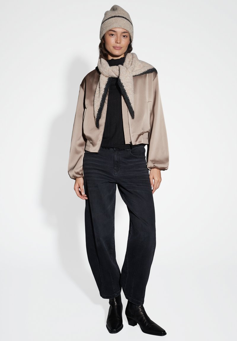 Giacca bomber cropped beige satin, top nero a costine, jeans neri a vita alta, sciarpa beige con bordo scuro e berretto lavorato a maglia beige.