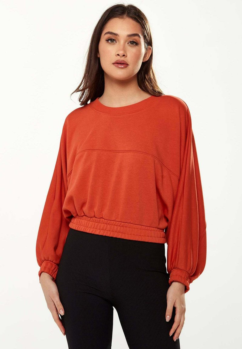 Orange pullover sweater with a relaxed fit, elasticized hem, and voluminous sleeves. Paired with black ribbed leggings.