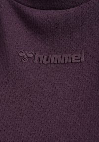 Fioletowa, teksturowana tkanina z małymi otworami, z wytłoczonym logo "hummel" i wzorem pszczoły, podkreślająca oddychający, sportowy materiał.