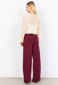 Frau mit langen blonden Haaren, die ein cremefarbenes Spitzenshirt mit langen Ärmeln und hochtaillierte, weit geschnittene weinrote Hose trägt, steht vor einem weißen Hintergrund.