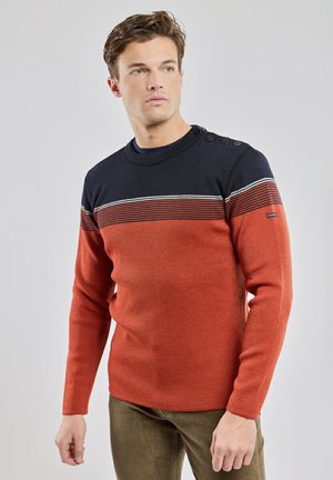 Ung mand iført en marineblå og brændt orange stribet sweater med knapper på skulderen og brune manchesterbukser, der kigger til siden.