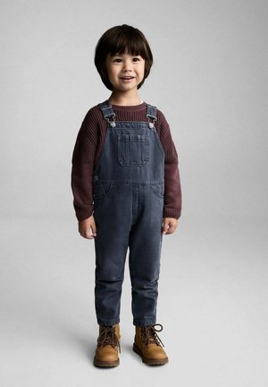 Salopette en denim bleu avec des bretelles ajustables, associée à un pull texturé bordeaux et des bottes à lacets marron, avec une poche avant.