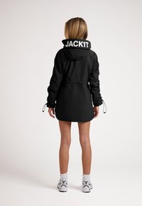 Giacca nera con cappuccio, vita cintrata e polsini elastici. Presenta grandi scritte bianche "JACK1T" sul colletto. Texture liscia.
