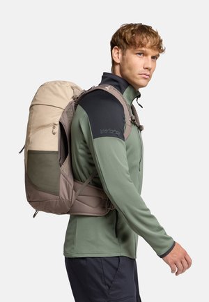 Sac à dos léger, beige et vert, avec des bretelles rembourrées, des accents en maille et des zips, porté sur un haut à manches longues vert, vue de côté.