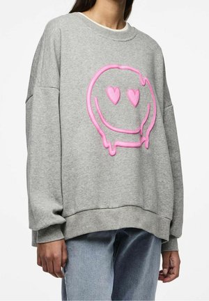 Personne portant un sweat-shirt gris ample avec un grand visage souriant rose qui fond, avec des yeux en forme de cœur, sur le devant.