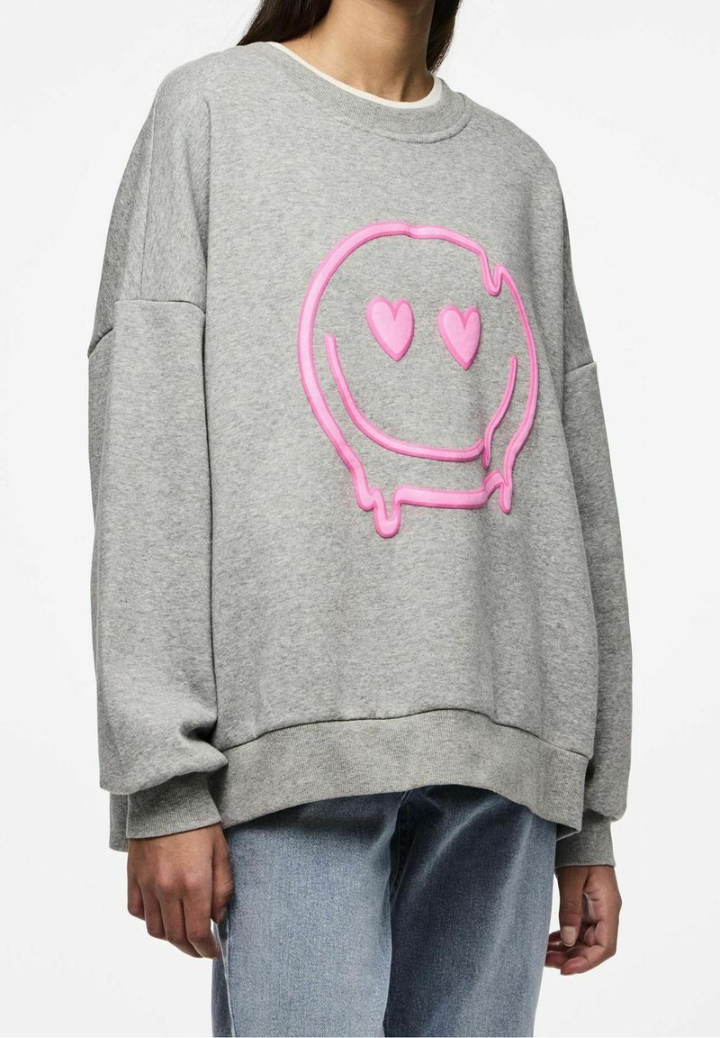 Personne portant un sweat-shirt gris ample avec un grand visage souriant rose qui fond, avec des yeux en forme de cœur, sur le devant.