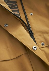 Veste marron à finition mate, dotée d'une fermeture éclair noire, de boutons-pression en argent et d'une doublure intérieure à motifs. Design simpliste avec des lignes épurées.