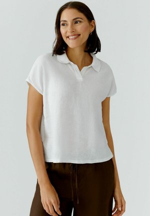 Frau mit schulterlangem dunklem Haar, die lächelt, trägt ein kurzärmeliges weißes Hemd mit Kragen und dunkelbraune Hosen vor einfachem Hintergrund.