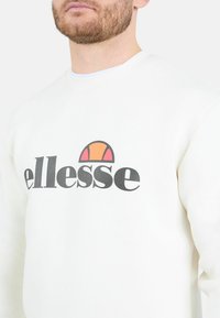 Sudadera de color crema hecha de una mezcla de algodón, con el logo "ellesse" en negro y un círculo de acento en color encima. Cuello de crew clásico.