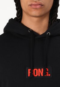 Black hoodie made of cotton, featuring a prominent red embroidered logo on a rectangular patch. Includes gray drawstring and eyelets.