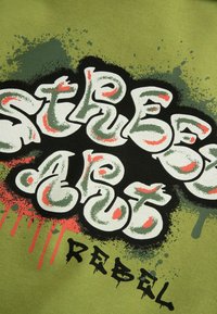 Zielony materiał zdobi odważny biały tekst w stylu graffiti, który brzmi "STREET ART" z czarnymi konturami i czerwonymi akcentami, obok słowa "REBEL".