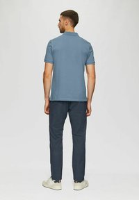 Mann steht mit dem Rücken zur Kamera, trägt ein blaues Kurzarm-Polohemd, dunkle Hosen und weiße Sneakers vor einem schlichten hellen Hintergrund.