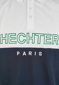 Witte polo met groene borduring van "HECHTER" en "PARIS" in het wit. Voorzien van drie knopen en een contrasterende marineblauwe onderpaneel. Gemaakt van katoen.