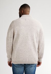 Světle šedý svetr s vysokým límcem, s texturovaným žebrovaným designem na ramenou a měkkým pleteným povrchem. Nosený s modrými džíny.