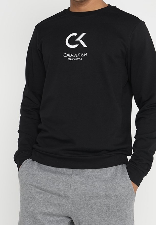 Sweat-shirt noir à manches longues avec un col rond, présentant un logo "CK" blanc et le texte "CALVIN KLEIN PERFORMANCE". Le tissu semble lisse et léger.
