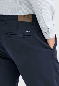 marineblå bomullsbukser har en baklomme med et lite brodert emblem i rødt, hvitt og blått. Merket sier "FQ1924 AUTHENTIC JEANS."