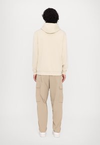 Felpa color crema con cappuccio e cordino, abbinata a pantaloni cargo beige con tasche laterali e caviglie affusolate, posizionata su uno sfondo semplice.