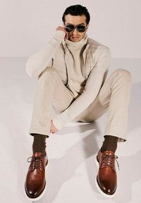 Scarpe eleganti in pelle marrone con suole bianche, indossate con pantaloni beige, calzini marroni e un dolcevita crema con giacca trapuntata; occhiali da sole scuri.