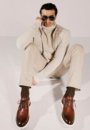 Scarpe eleganti in pelle marrone con suole bianche, indossate con pantaloni beige, calzini marroni e un dolcevita crema con giacca trapuntata; occhiali da sole scuri.