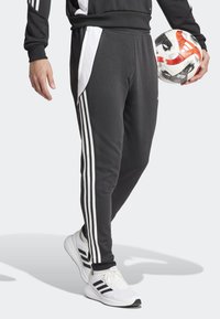 Schwarze Sporthose mit weißen Seitenpanelen und drei vertikalen Streifen. Getragen mit weißen Turnschuhen und einem gemusterten Fußball in der Hand.