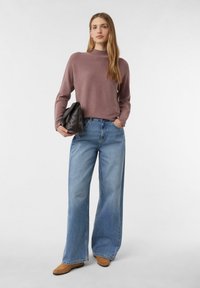 Brauner Strickpullover mit hohem Kragen, kombiniert mit weit geschnittenen hellblauen Jeans und hellbraunen Schuhen. Hält eine schwarze strukturierte Tasche.