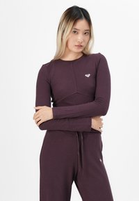 Mladá žena s dlhými blond vlasmi, oblečená v zladenom tmavo fialovom rebrovanom topi s dlhým rukávom a nohaviciach, s prekríženými rukami na hrudi.