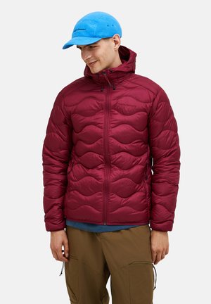 Man wearing a maroon quilted jacket with a hood, blue fleece cap, and brown cargo pants, looking down and smiling.