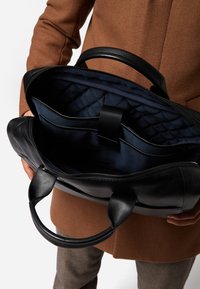Schwarze Lederhandtasche mit Reißverschluss, ausgestattet mit einem gesteppten navyblauen Innenraum und einer Organisationsasche. Dunkle Griffe und eine strukturierte Form.