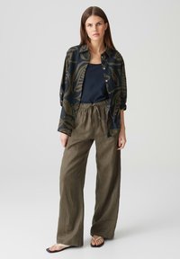 Pantaloni in lino verde oliva a gamba larga con vita elastica, abbinati a una maglietta senza maniche blu navy e una camicia a bottoni nera e verde a fantasia.