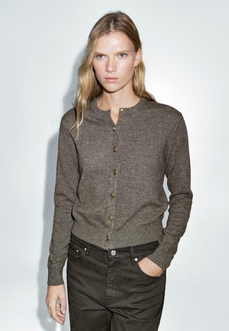 Cardigan in maglia marrone con scollatura tonda, maniche lunghe e bottoni dorati. Indossato sopra pantaloni scuri, con una vestibilità slim.