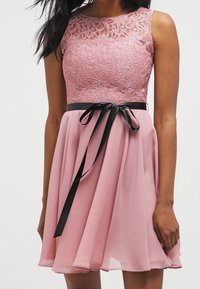 Robe rose avec un corsage en dentelle et une jupe fluide. Comprend une ceinture en ruban noir nouée à la taille pour accentuer.