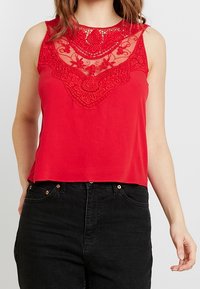Top senza maniche rosso con dettagli in pizzo sul collo e ricami floreali. Tessuto morbido con una vestibilità ampia, abbinato a jeans neri.