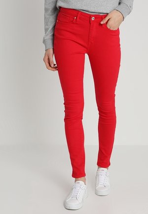 Persoon draagt felrode skinny jeans, grijze longsleeve en witte sneakers, met één hand in de zak, staand op een effen achtergrond.