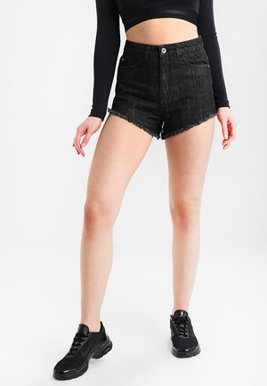 Γυναίκα που φοράει μαύρα σορτς τζιν με ψηλή μέση και ξεφτισμένα άκρα, μαύρο κοντομάνικο μπλουζάκι με μακριά μανίκια και μαύρα αθλητικά παπούτσια, στέκεται σε λευκό φόντο.