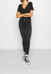Woman standing with arms crossed wearing black V-neck T-shirt, dark grey skinny jeans with knee seams, white socks, and black high-top sneakers.