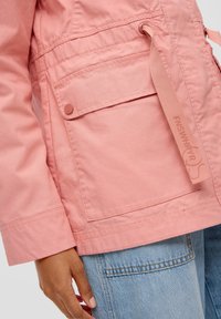 Detail ruky u světlé růžové bundy s klopou a knoflíkem, nošené přes modré džínové kalhoty.