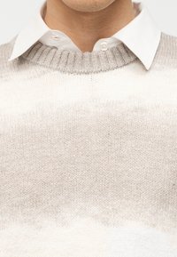 Gros plan sur une personne portant une chemise blanche à col sous un pull tricoté beige avec un col côtelé.