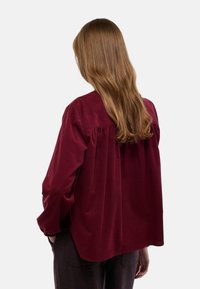 Chemise en velours côtelé bordeaux à coupe décontractée, avec un détail de dos froncé et des manches longues, montrée de dos.