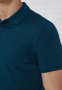 Mørk teal polo-skjorte med krage, korte ermer og en teksturert stoff med en glatt overflate. To knapper synlige ved halsåpningen.