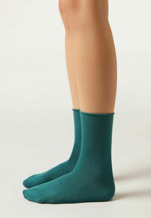 Piernas que llevan calcetas de color verde azulado sobre un fondo neutro, mostrando el ajuste de las calcetas y su suave textura.