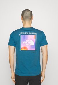 Korta ärmar teal t-shirt i mjukt tyg med en färgglad grafik på baksidan som visar "The North Face"-logotypen och textöverlagring.