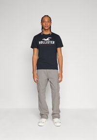 Melnas kokvilnas T-krekls ar "HOLLISTER" logo baltā un sarkanā krāsā, apvienots ar gaiši pelēkiem krampju biksēm un baltām kedām.