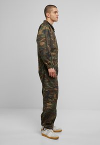 Zöld és barna árnyalatú camouflázs overál, elasztikus mandzsettákkal és elülső cipzárral, fehér és bézs sportcipőkkel párosítva.